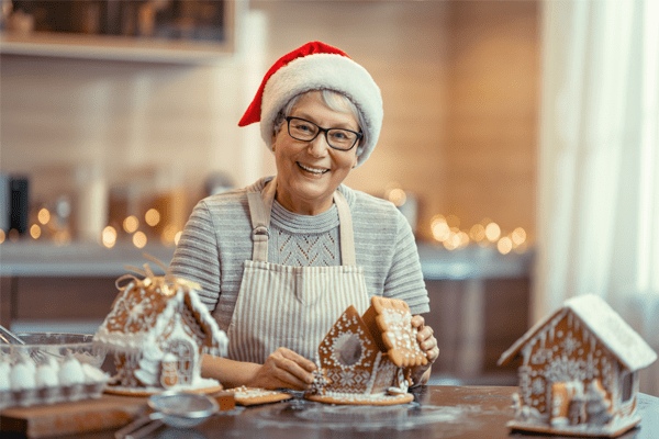 An older woman wearing a Santa hat and apron smiles while decorating gingerbread houses in a festive kitchen at MorningStar Senior Living Hillsboro. Holiday lights glow softly, creating a warm retirement living atmosphere.