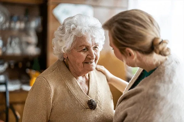 An elderly woman with white hair smiles warmly at a younger woman who gently touches her shoulder. They appear to be having a friendly conversation in the cozy, welcoming atmosphere of MorningStar Senior Living Hillsboro.