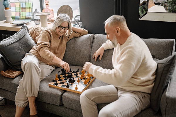 An older woman and man sit on a gray sofa, smiling and playing chess together in the cozy, warmly lit living room of MorningStar Senior Living Hillsboro—enjoying the comforts of retirement living.