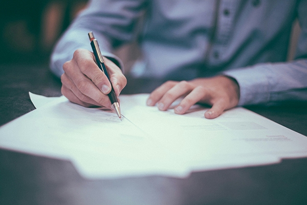 A person in a blue shirt writes on white sheets of paper with a black and gold pen while resting their other hand on the documents, capturing a quiet moment at MorningStar Senior Living Hillsboro. The focus is on the hands and pen.