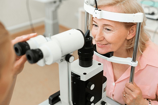 An older woman smiles while sitting at a slit lamp during an eye exam at MorningStar Senior Living Hillsboro, as an optometrist prepares to check her eyes in a comfortable retirement living setting.