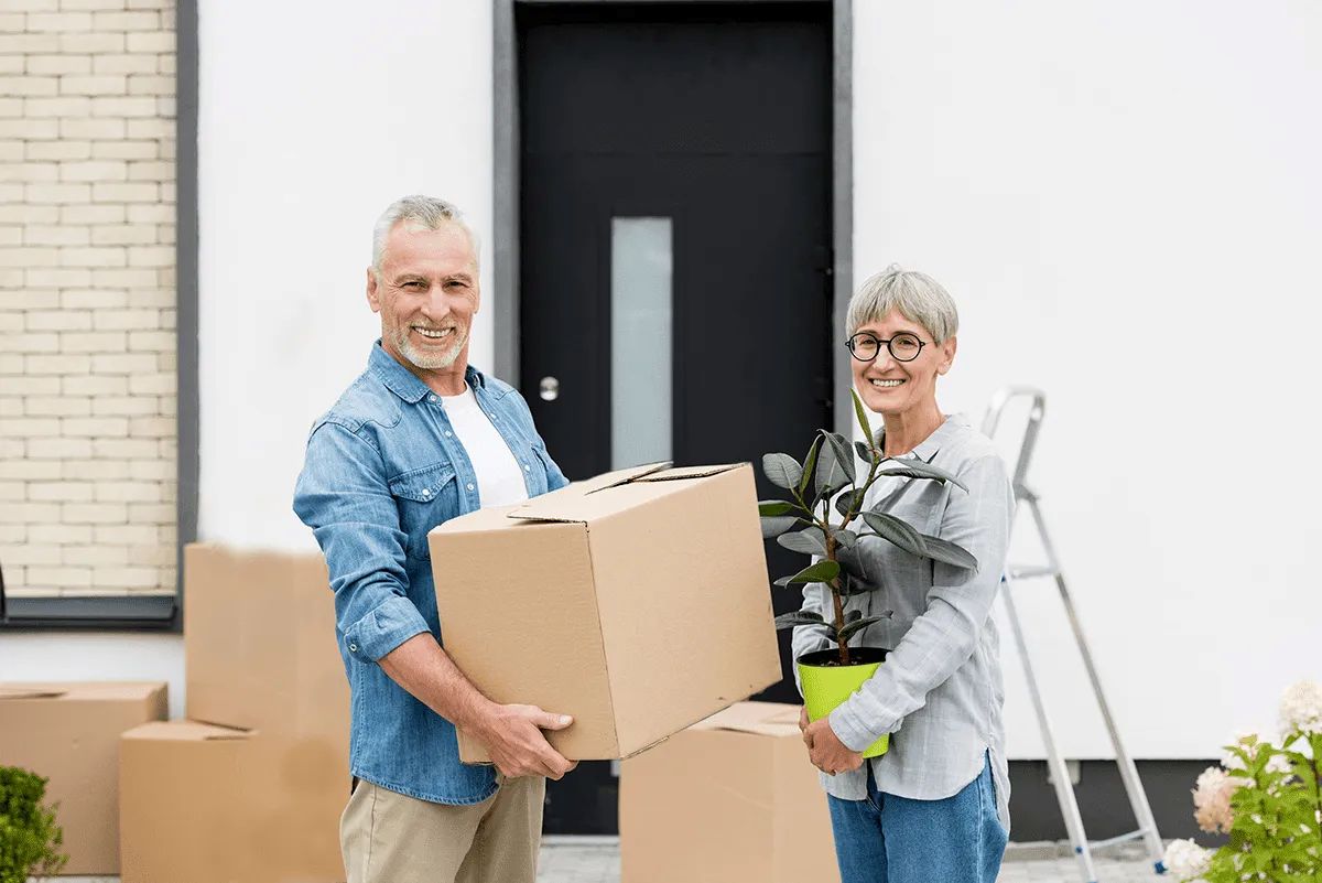 An older man and woman stand outside a house, smiling. The man holds a cardboard box, and the woman holds a potted plant. Several boxes and a ladder in the background suggest they are moving into Retirement Living at MorningStar Senior Living Hillsboro.