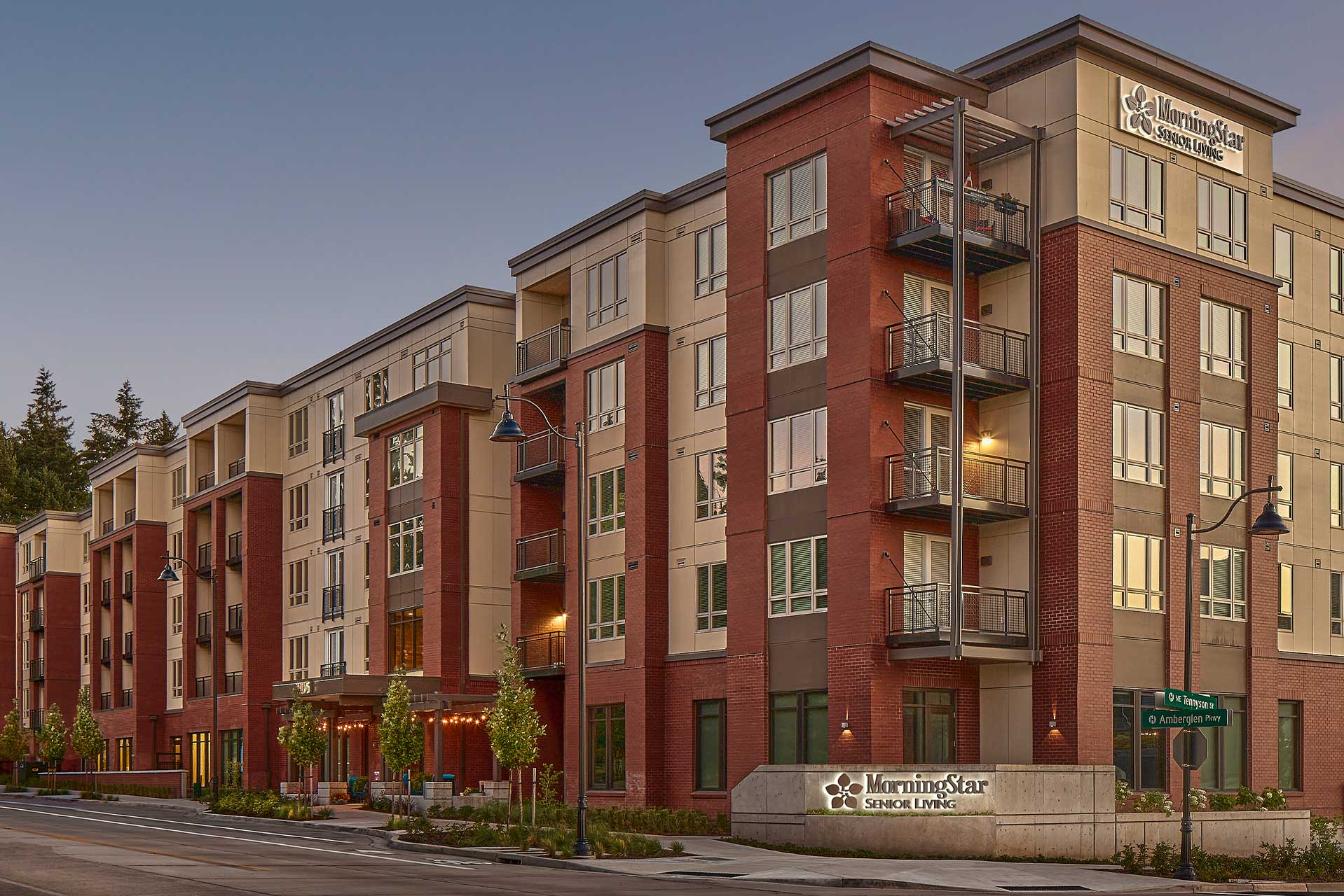 A modern, multi-story building with red brick and beige panels, featuring balconies and large windows. A sign in front reads