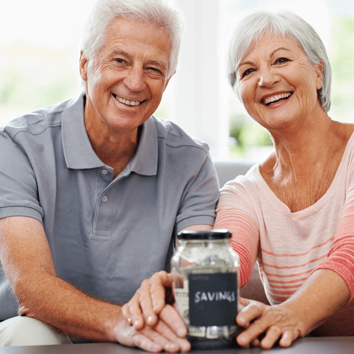 Smiling older couple sitting together at MorningStar Senior Living Hillsboro, holding a glass jar labeled "savings" filled with cash, their hands touching the jar, symbolizing retirement living and careful financial planning.