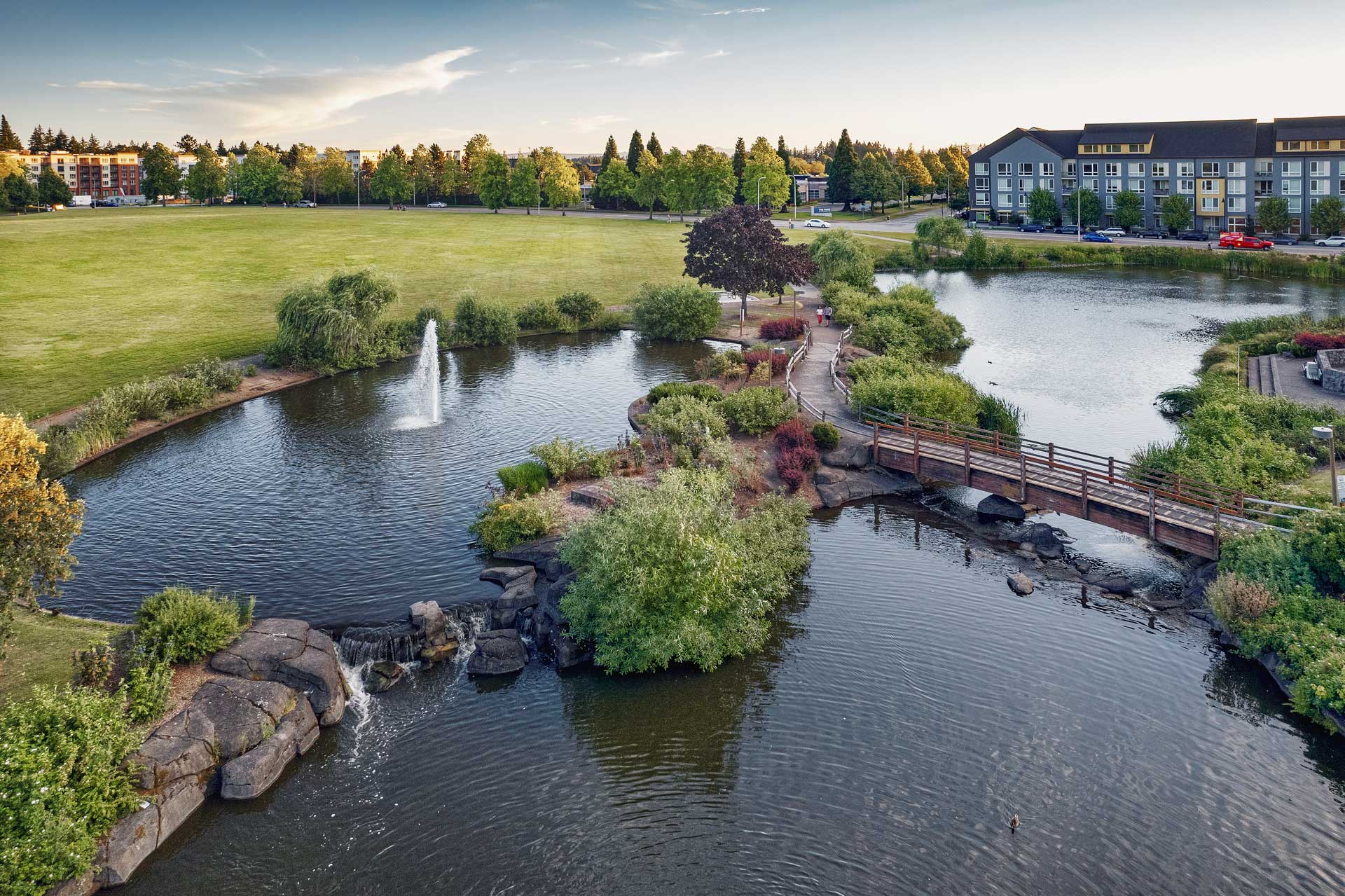 A park with a pond, footbridge, small waterfall, water fountain, and green lawns. Several buildings and trees surround the park under a clear sky—an inviting space near MorningStar Senior Living Hillsboro for relaxation and retirement living.