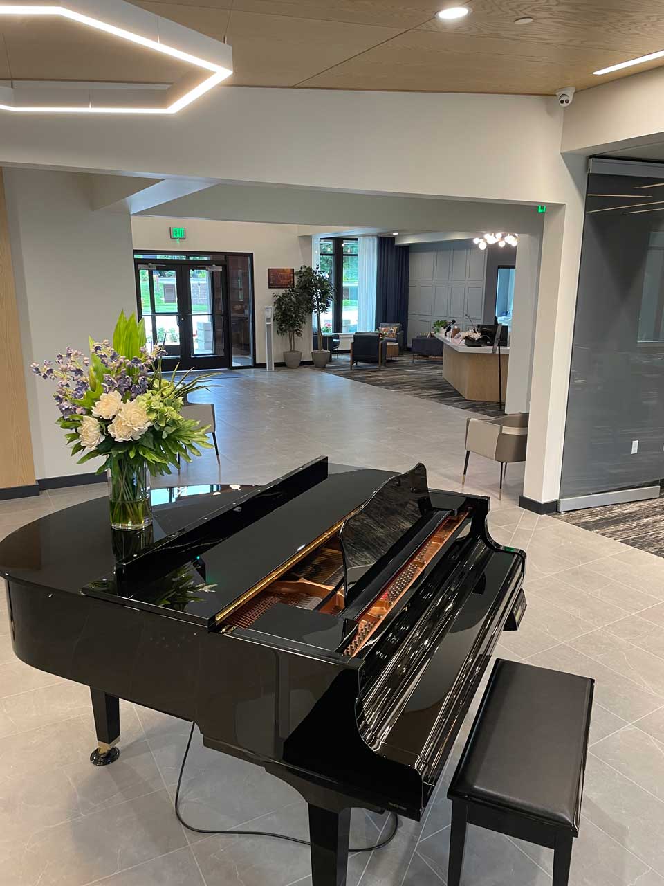 A grand piano with a vase of fresh flowers sits in the modern, spacious lobby of MorningStar Senior Living Hillsboro, featuring gray tile floors, contemporary furniture, and large windows that fill this retirement living space with natural light.