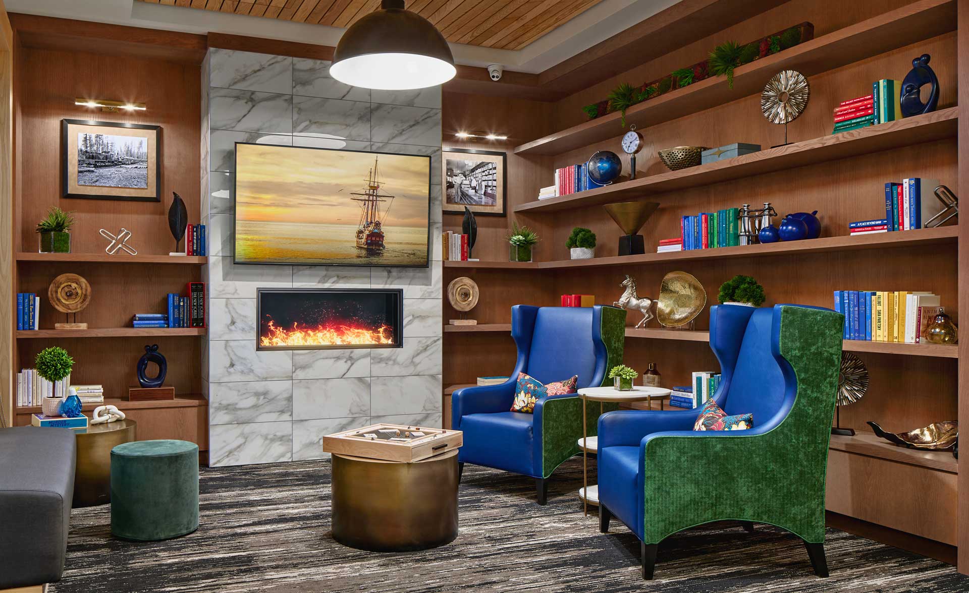 Modern library lounge at MorningStar Senior Living Hillsboro features wooden shelves filled with books and decor, colorful armchairs, a gold coffee table, ottomans, wall art, and a marble fireplace with a TV displaying a ship above it.