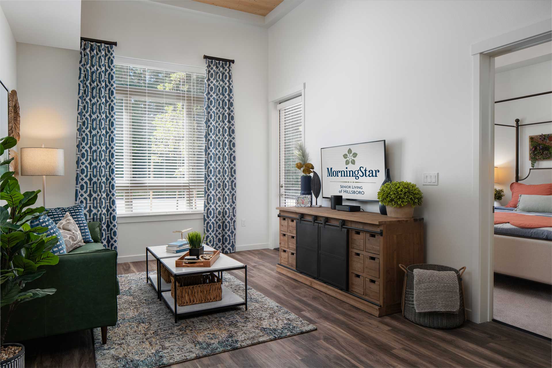 Modern living room with a green sofa, patterned rug, large windows with blue curtains, TV displaying