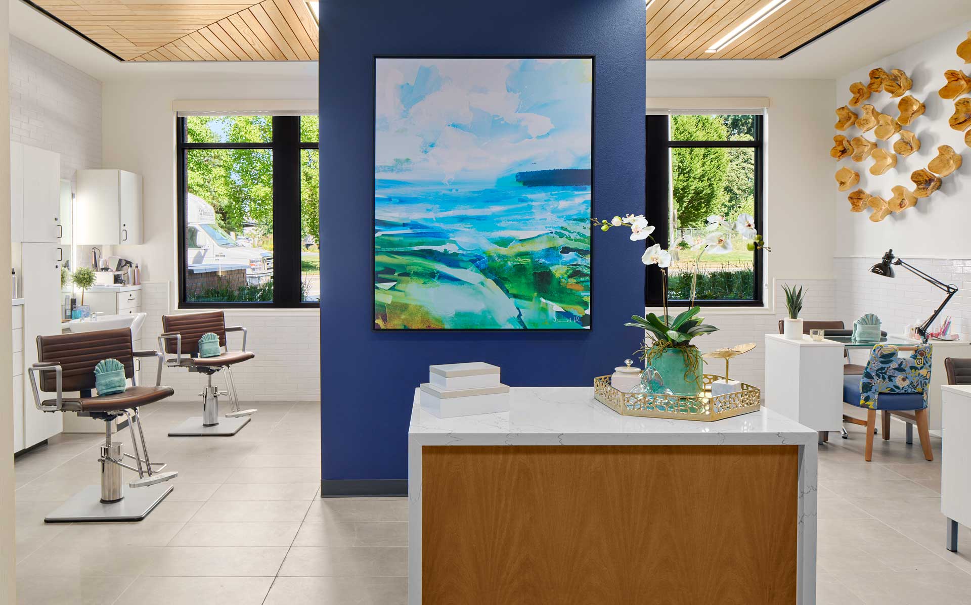 Modern salon interior at MorningStar Senior Living Hillsboro with a reception desk in front, a blue accent wall with vibrant abstract art, styling chairs to the left, large windows, and bright natural light—ideal for elegant retirement living.