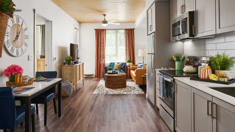 Modern apartment ideal for Retirement Living, featuring an open kitchen with gray cabinets and stainless steel appliances. The living area boasts an orange sofa, round coffee table, and large windows—perfect for comfortable living at MorningStar Senior Living Hillsboro.
