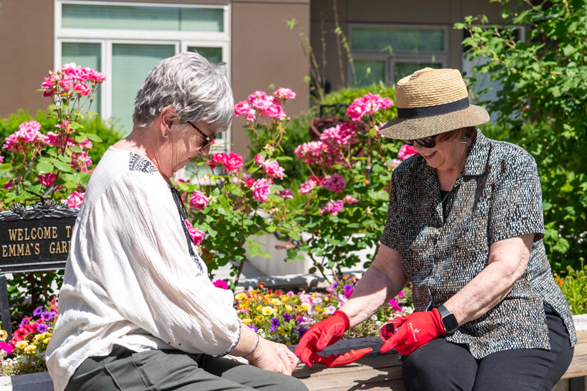 Two older women sit on a bench in a garden full of colorful flowers at MorningStar Senior Living Hillsboro. One wears a straw hat and red gardening gloves, while the other, with gray hair and glasses, enjoys the sunny day and retirement living.