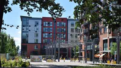 Modern apartment buildings with large windows surround a pedestrian plaza, where residents of MorningStar Senior Living Hillsboro enjoy outdoor seating, greenery, string lights, and a vibrant retirement living community on sunny days.