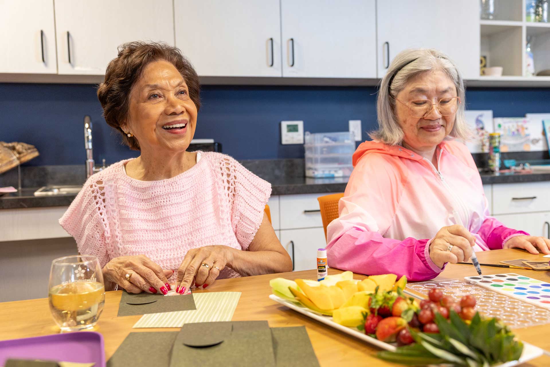 Two older women enjoy arts and crafts at MorningStar Senior Living Hillsboro. One smiles while folding paper; the other draws with a marker. A plate of fruit and cheese sits before them in a bright, cheerful retirement living space.