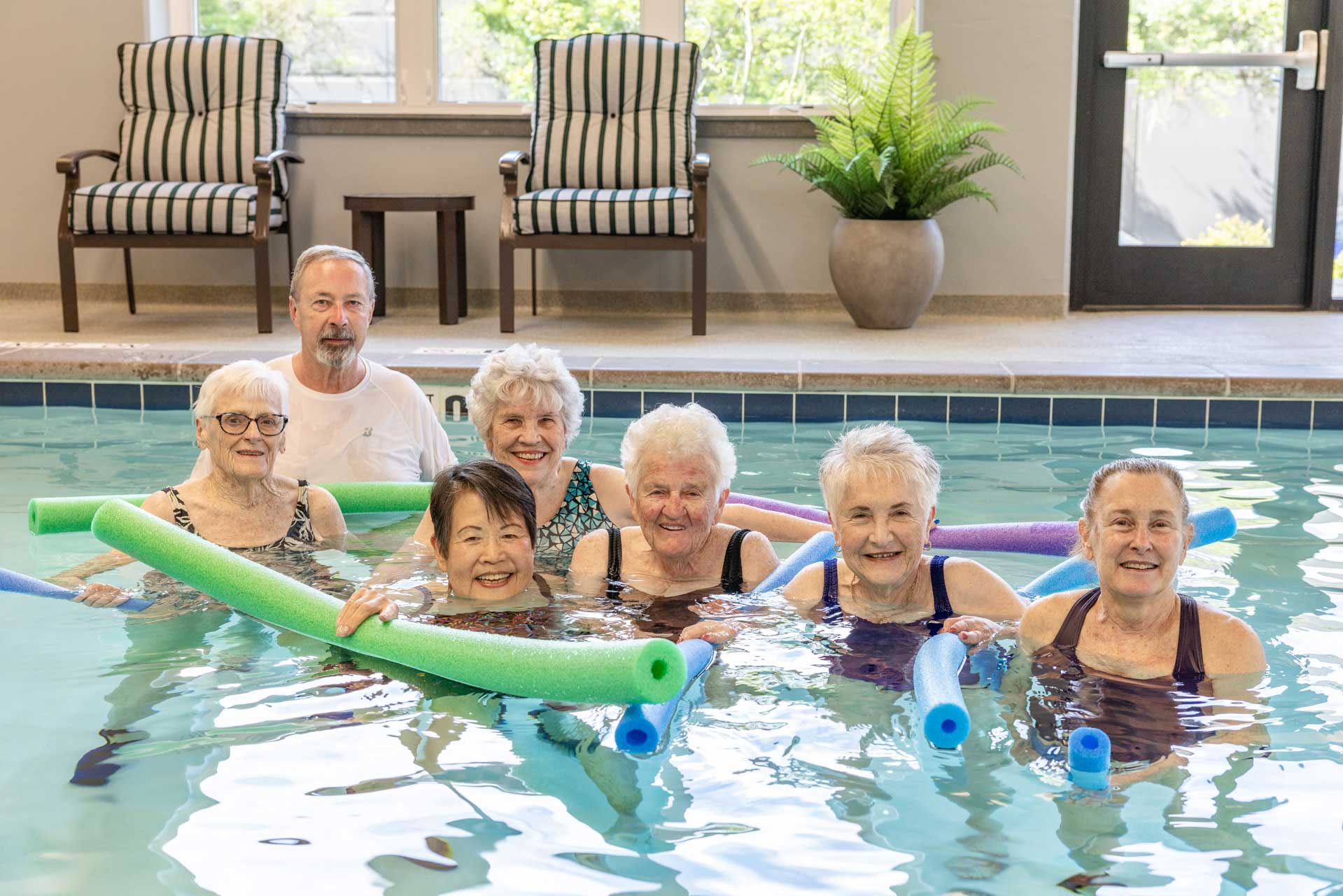 A group of six older women and one older man smile while standing in a pool at MorningStar Senior Living Hillsboro, holding colorful pool noodles, with chairs, a plant, and windows visible in the background—enjoying vibrant retirement living.