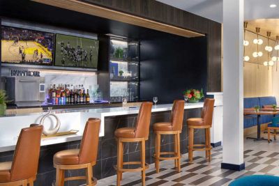 A modern bar at MorningStar Senior Living Hillsboro features brown leather barstools, a white counter, and liquor bottles on shelves. Two TVs display basketball and football games. The checkered floor leads to a cozy retirement living seating area.