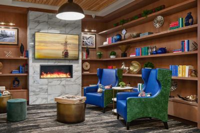 A modern library lounge at MorningStar Senior Living Hillsboro features wooden shelves, colorful books, two blue and green armchairs, a round ottoman, a small table, a lit fireplace, and a TV above it—perfect for relaxed retirement living.