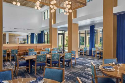 Modern restaurant dining room at MorningStar Senior Living Hillsboro with blue upholstered chairs, round and square wooden tables set with plates and glasses, large windows, blue curtains, and hanging lights—a bright, spacious setting for Retirement Living.