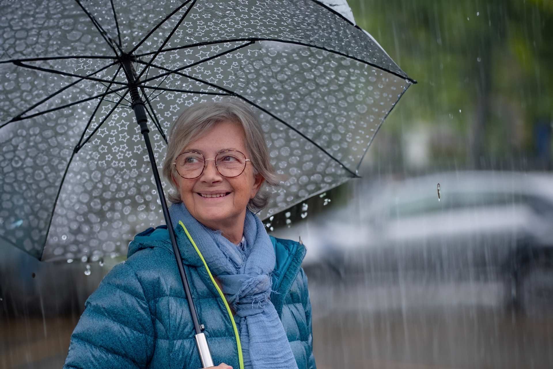 smiling-senior-woman-walking-in-the-street-in-the-rain A smiling senior woman standing under an umbrella in the rain.
